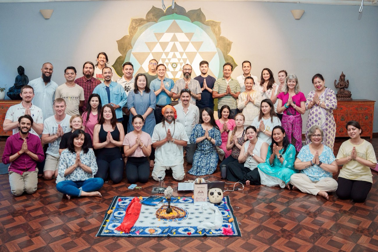Eine große Gruppe von Menschen mit Händen im Gebetsgruß nach einer Meditation, vor einem Sri-Yantra-Wandbild